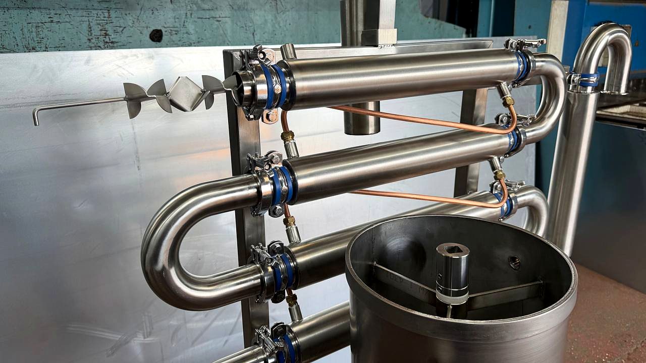 Coiled stainless pipework with blue clamps on a stainless frame, beside a stainless tank