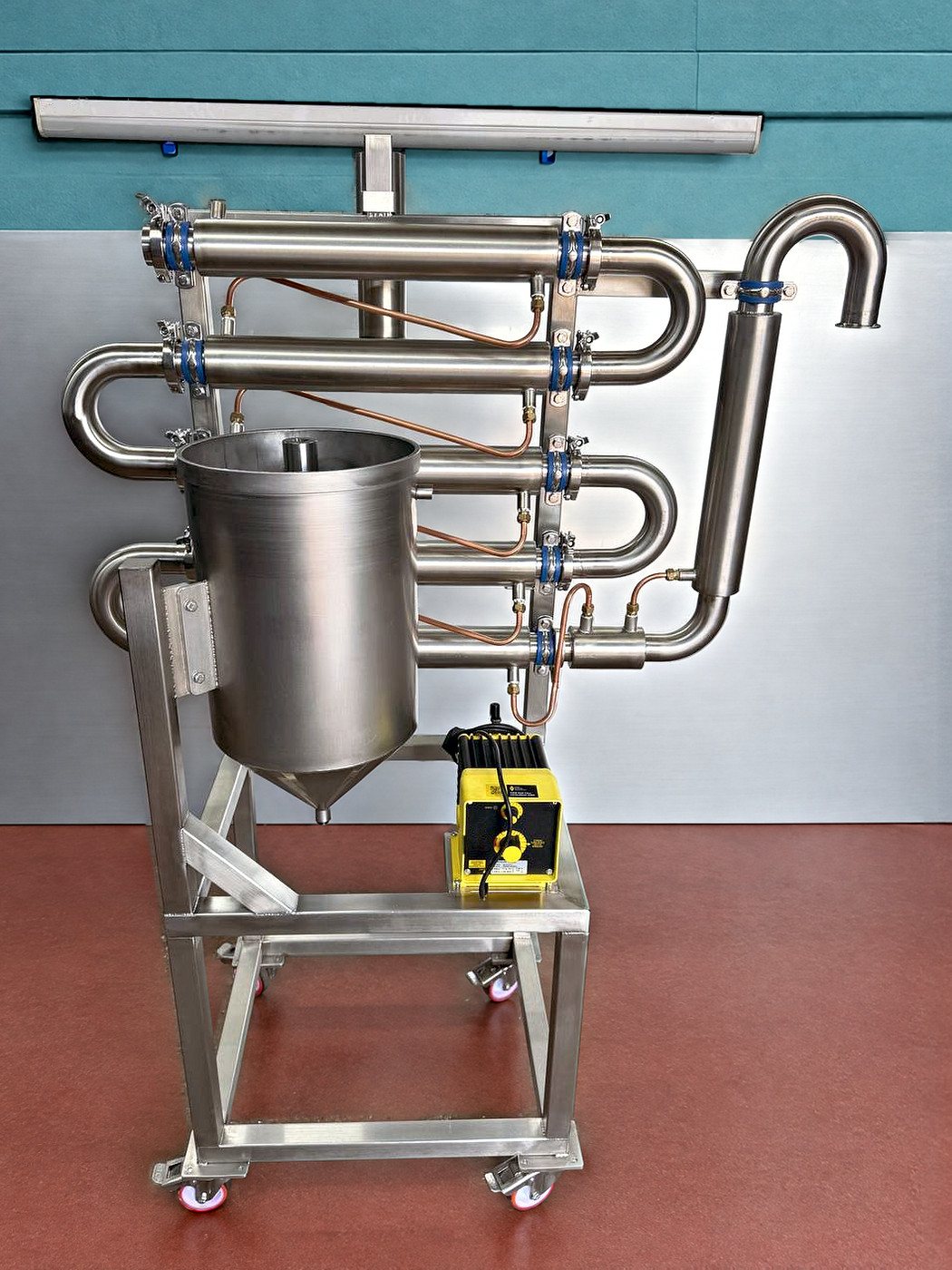 A stainless steel frame on castors carrying coiled stainless pipework, a conical tank and a yellow pump