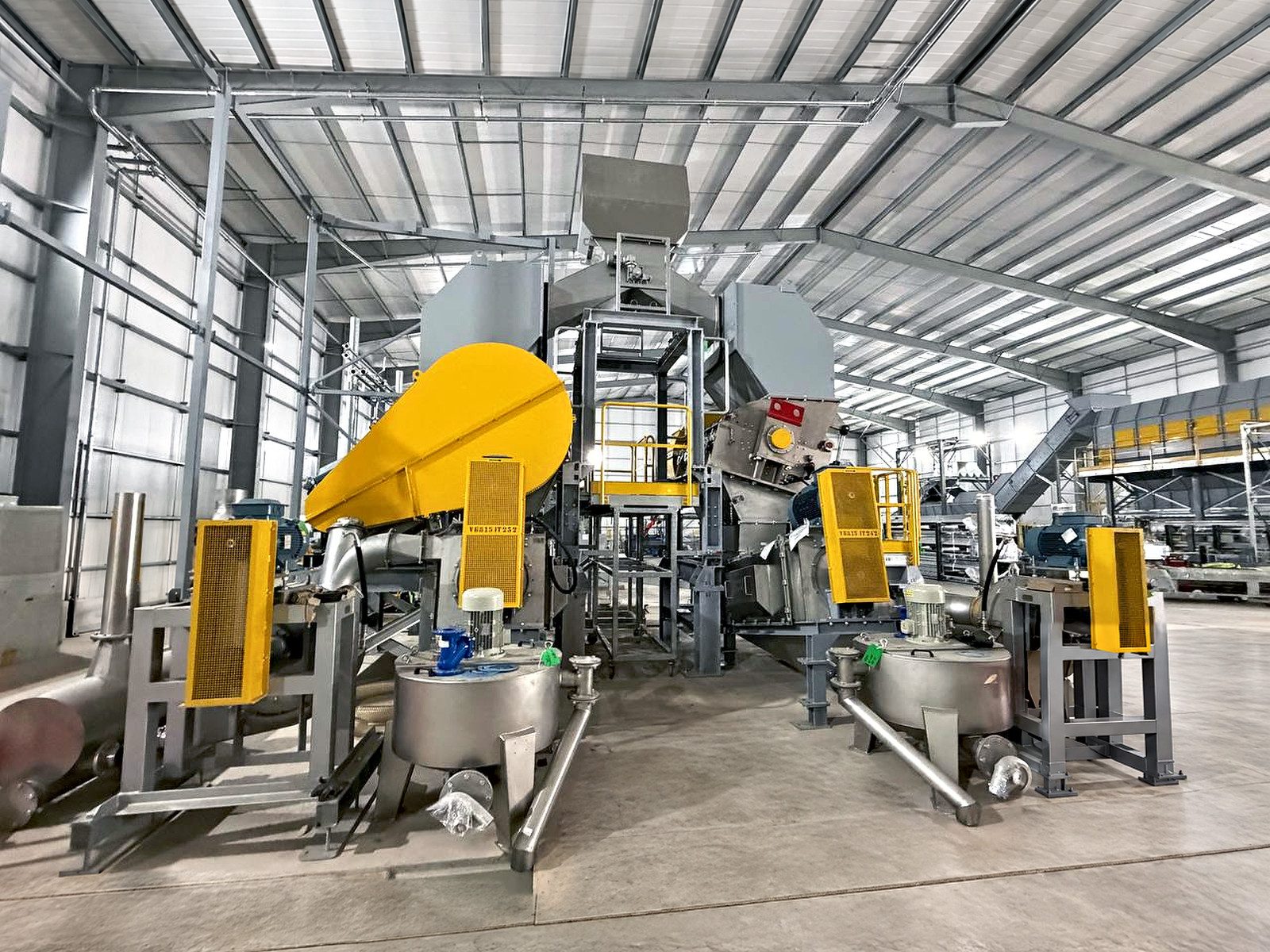 A processing line with a yellow hopper, stainless tanks and yellow mesh guards in a large factory