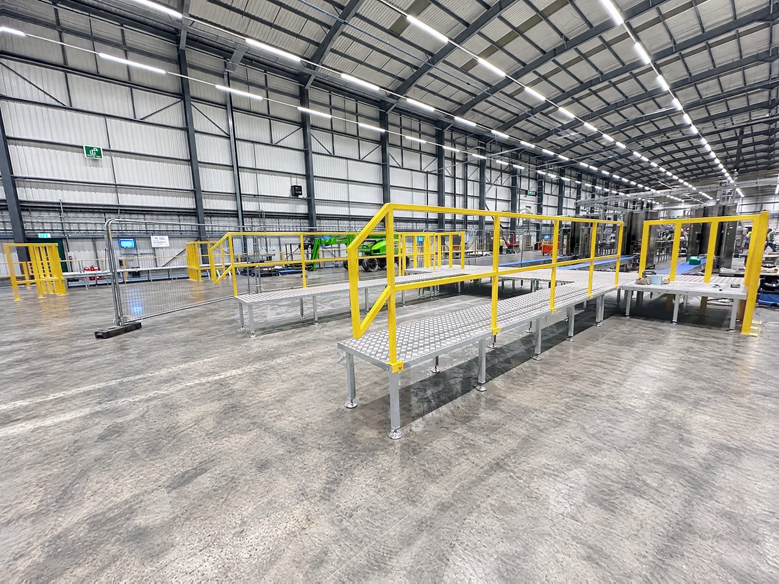 The corner of a raised galvanised walkway with yellow handrails, more walkway sections and gates behind it, in a warehouse