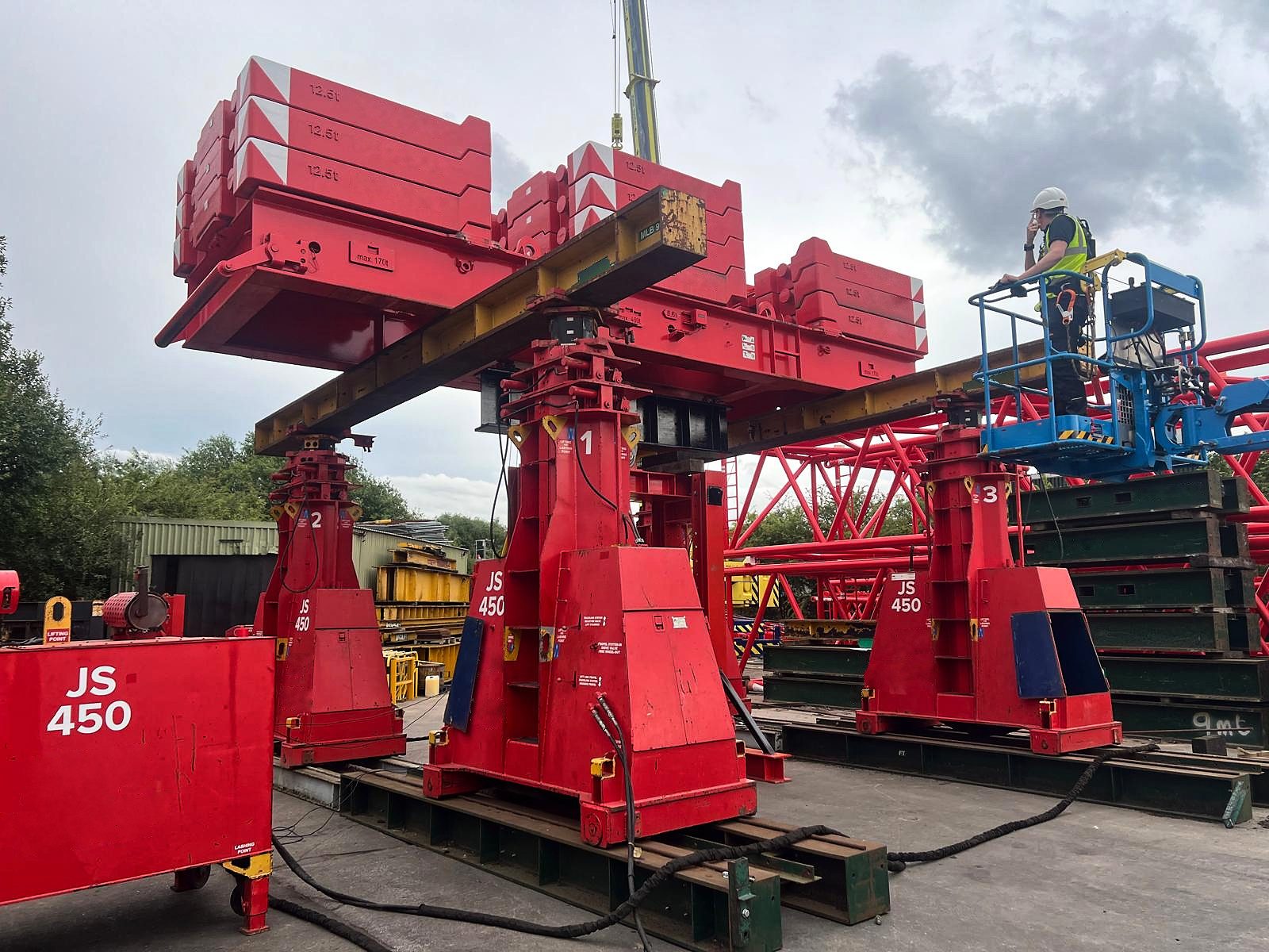 Three red jacking towers holding a yellow steel beam under stacks of red counterweights, with a worker in a blue work platform on the right