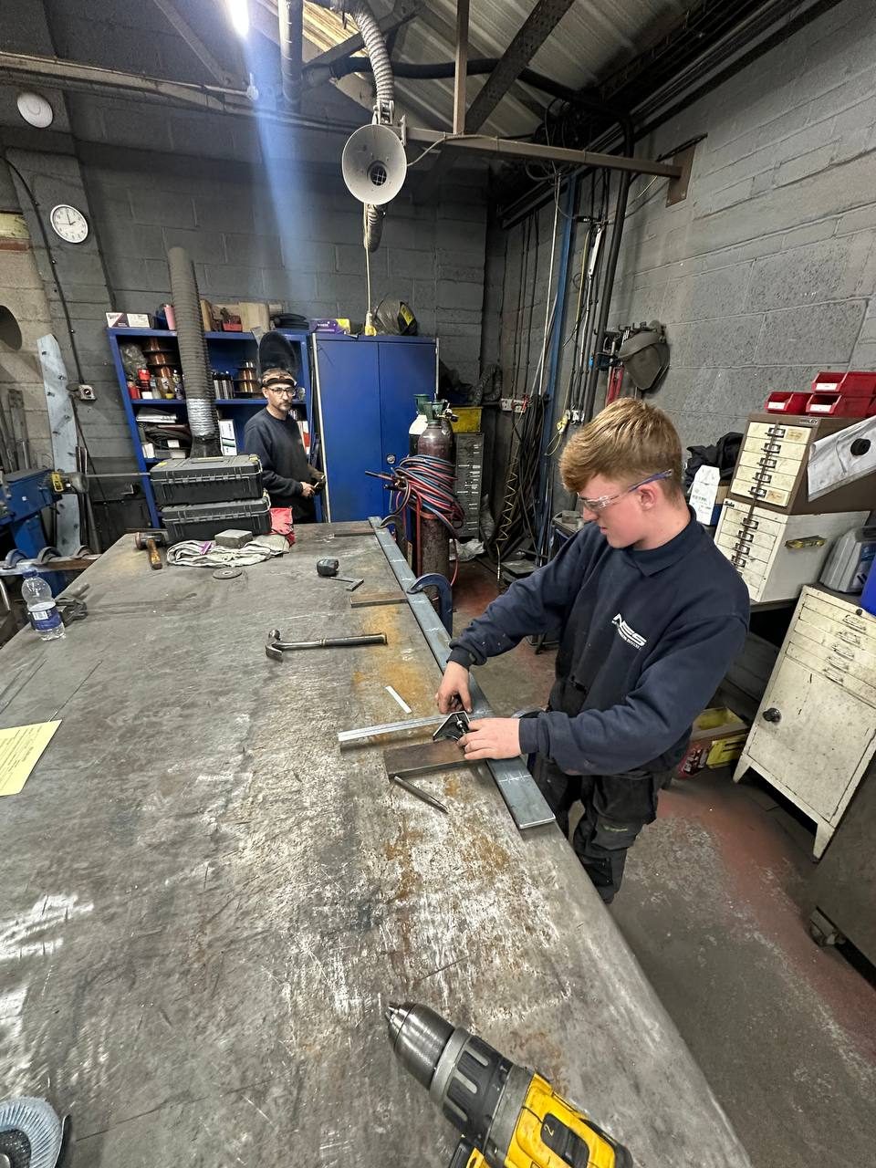 Marking out a steel bar with a combination square on the workshop bench, safety glasses on, the NES mark on the sweatshirt