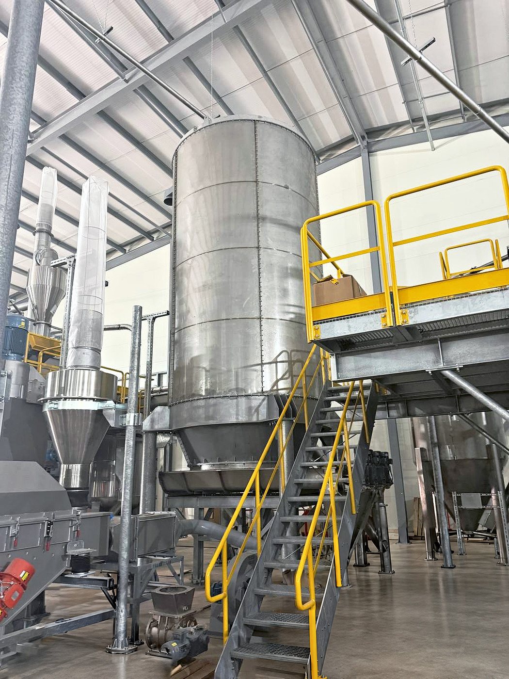 A stainless steel silo with a yellow handrailed platform and a galvanised staircase, inside a factory
