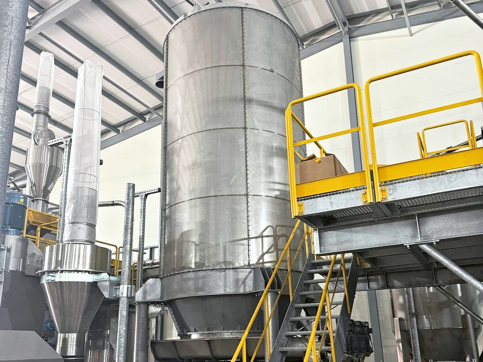 A stainless steel silo with a yellow handrailed platform and the top of a galvanised staircase, inside a factory