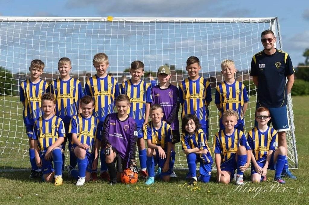 A junior football team in blue and yellow striped kit, the keepers in purple, NES across the front of the shirts, lined up in front of a goal with a man in the team colours at the end of the back row