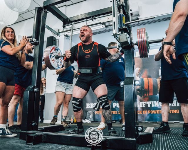 Jon May under a loaded bar in a squat rack on a lifting platform, spotters either side