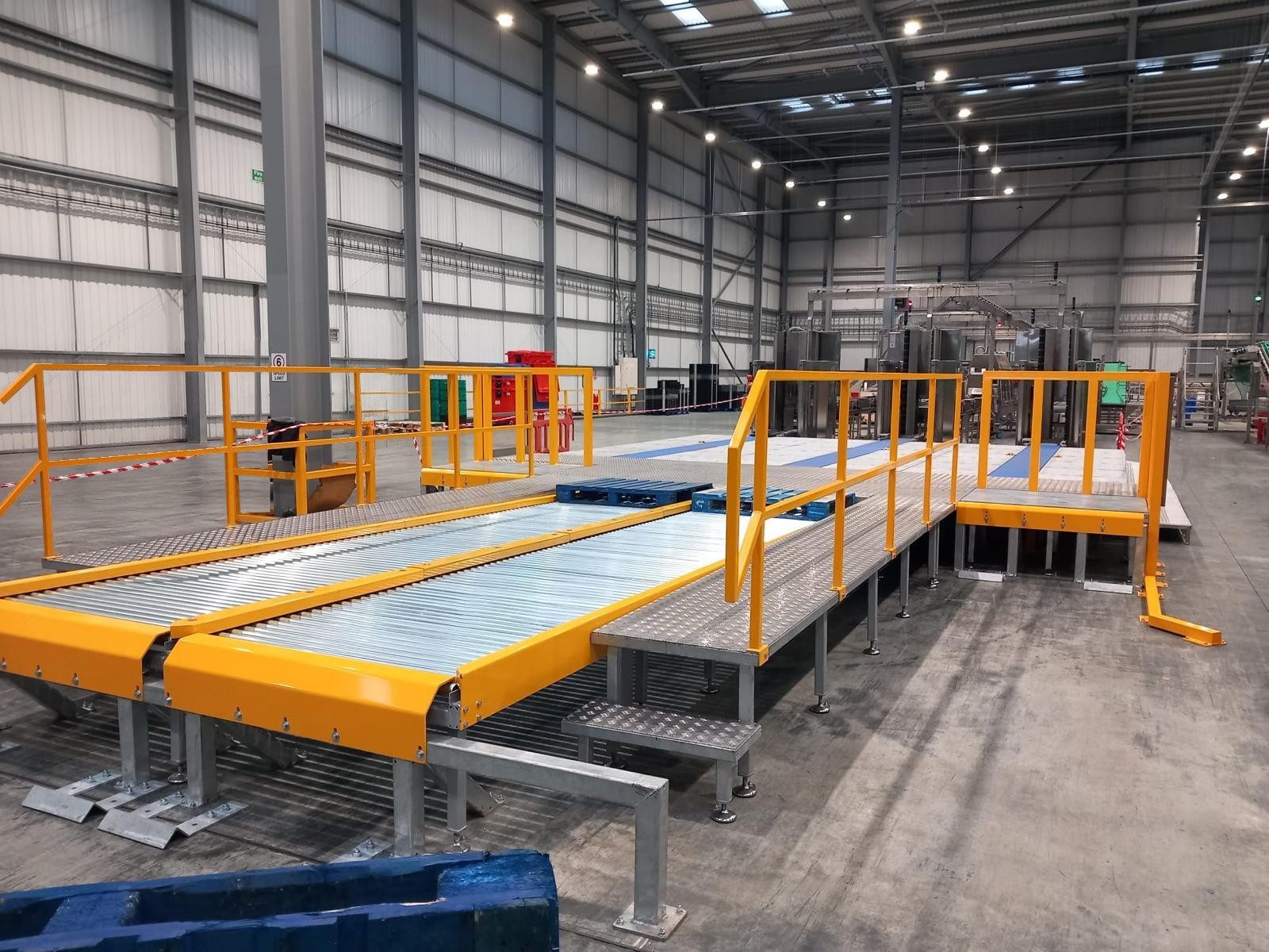 Galvanised roller bed conveyors with yellow guards beside a raised chequer-plate walkway with yellow handrails and a step, inside a large warehouse